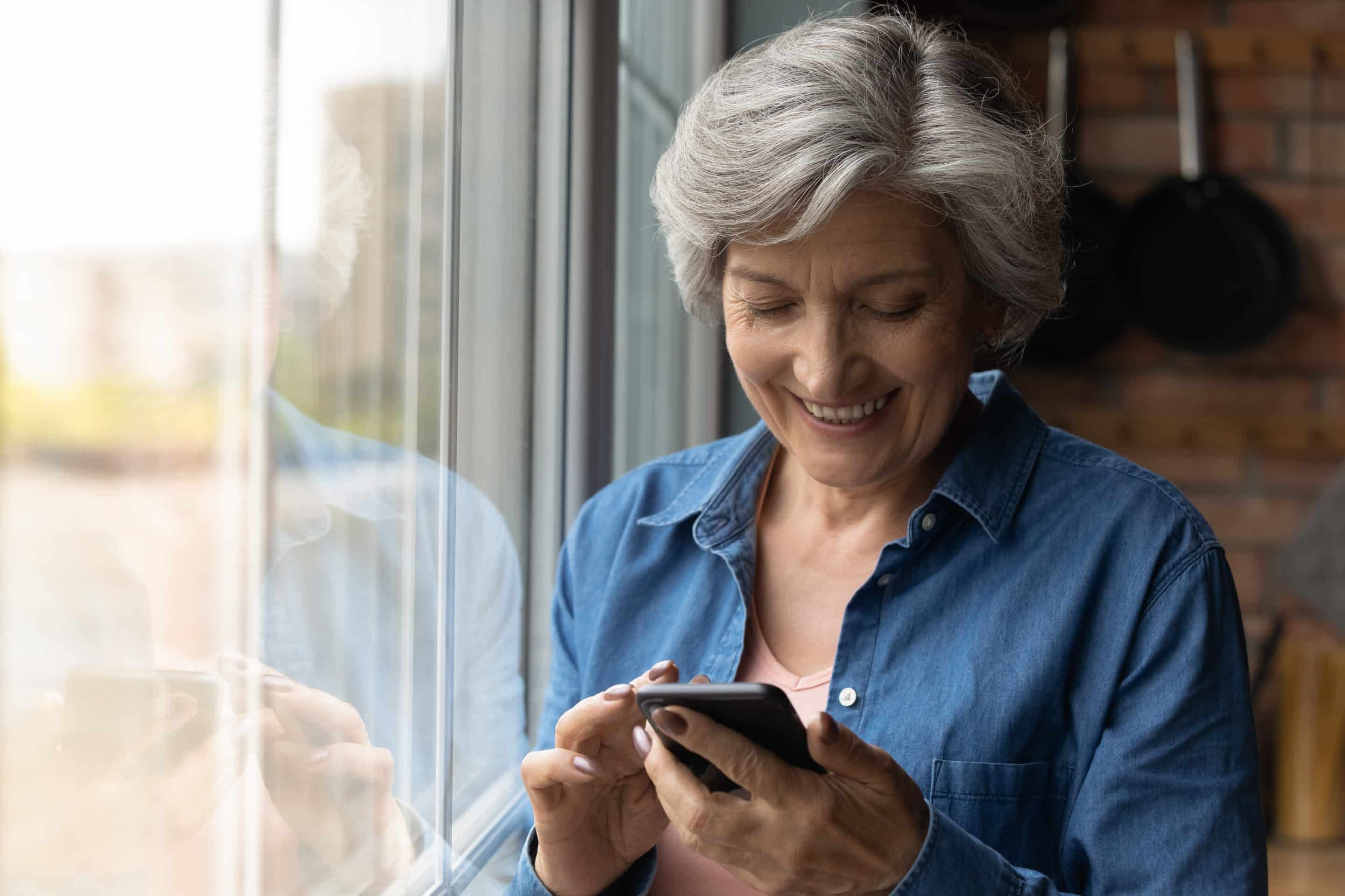 An older lady in a blue shirt smiling down at her phone by the window